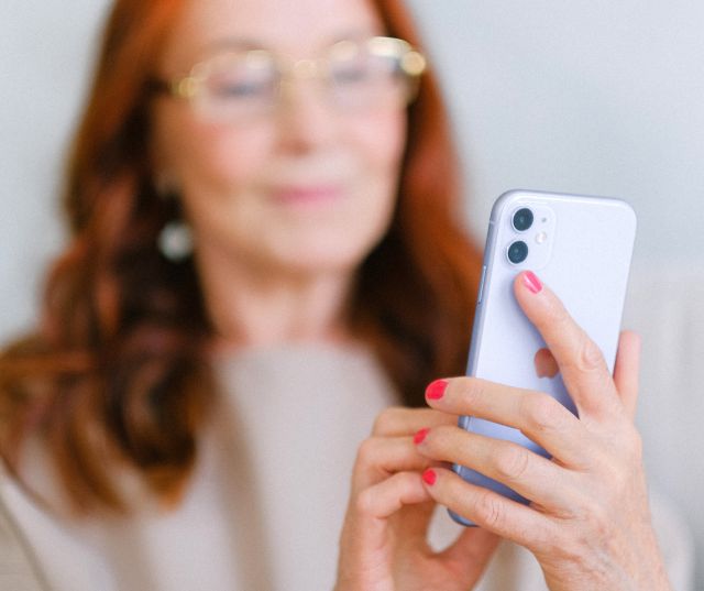 Crop aged redhead female in eyeglasses using contemporary mobile phone while surfing internet in soft focus