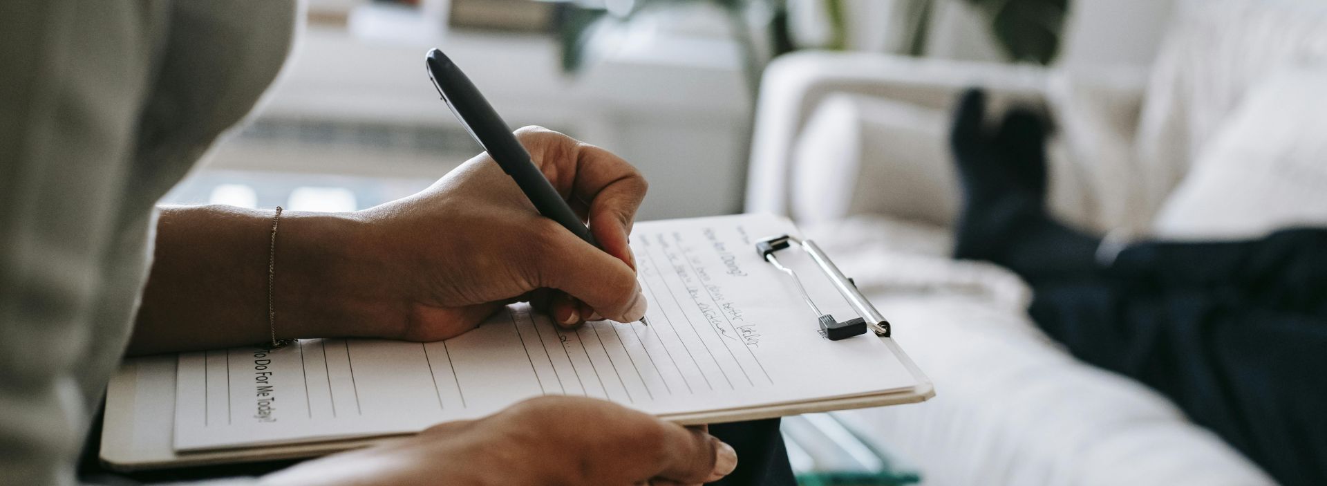 Unrecognizable ethnic female therapist taking notes on clipboard while filling out form during psychological appointment with anonymous client lying on blurred background